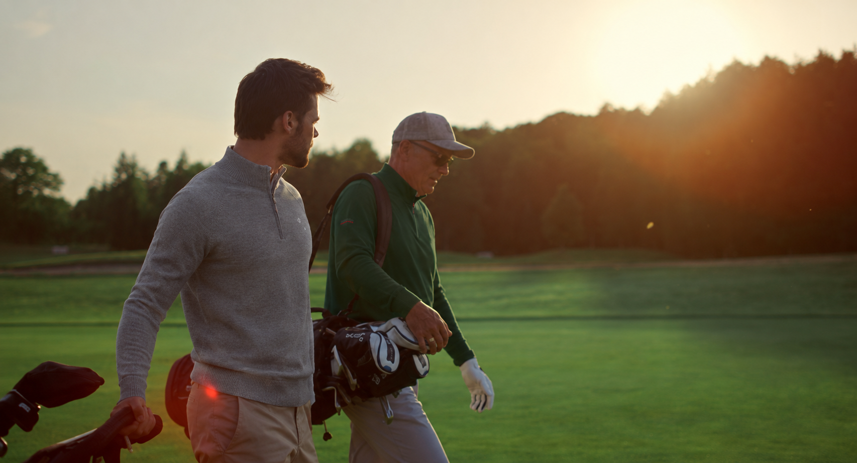 Two golfers walking a fairway at golden hour carrying bags, in grey and green quarter-zip layers
