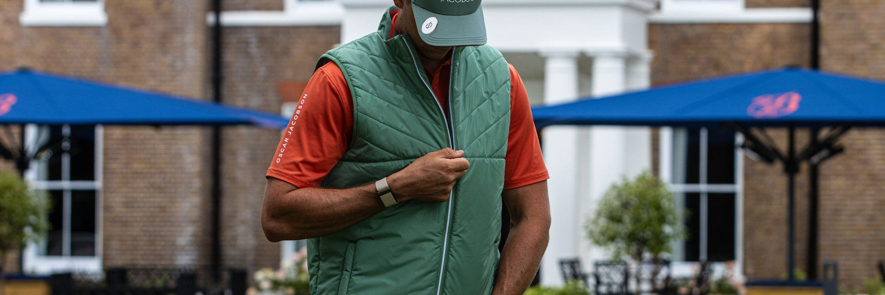 A golfer zipping a green Oscar Jacobson gilet over an orange polo outside a clubhouse